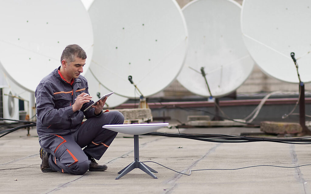 Um técnico de uniforme cinza está ajoelhado em um telhado, usando um tablet ao lado de uma antena parabólica, com diversas antenas parabólicas ao fundo