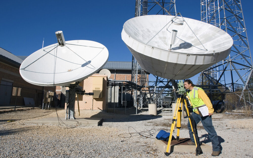 A technician in a yellow safety vest and blue jeans is using surveying equipment near two large satellite dishes at a communications site