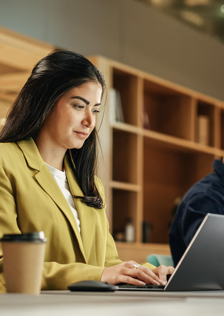 A businesswoman in a yellow blazer is working on a laptop in a modern office, with a coffee cup next to her and shelves in the background