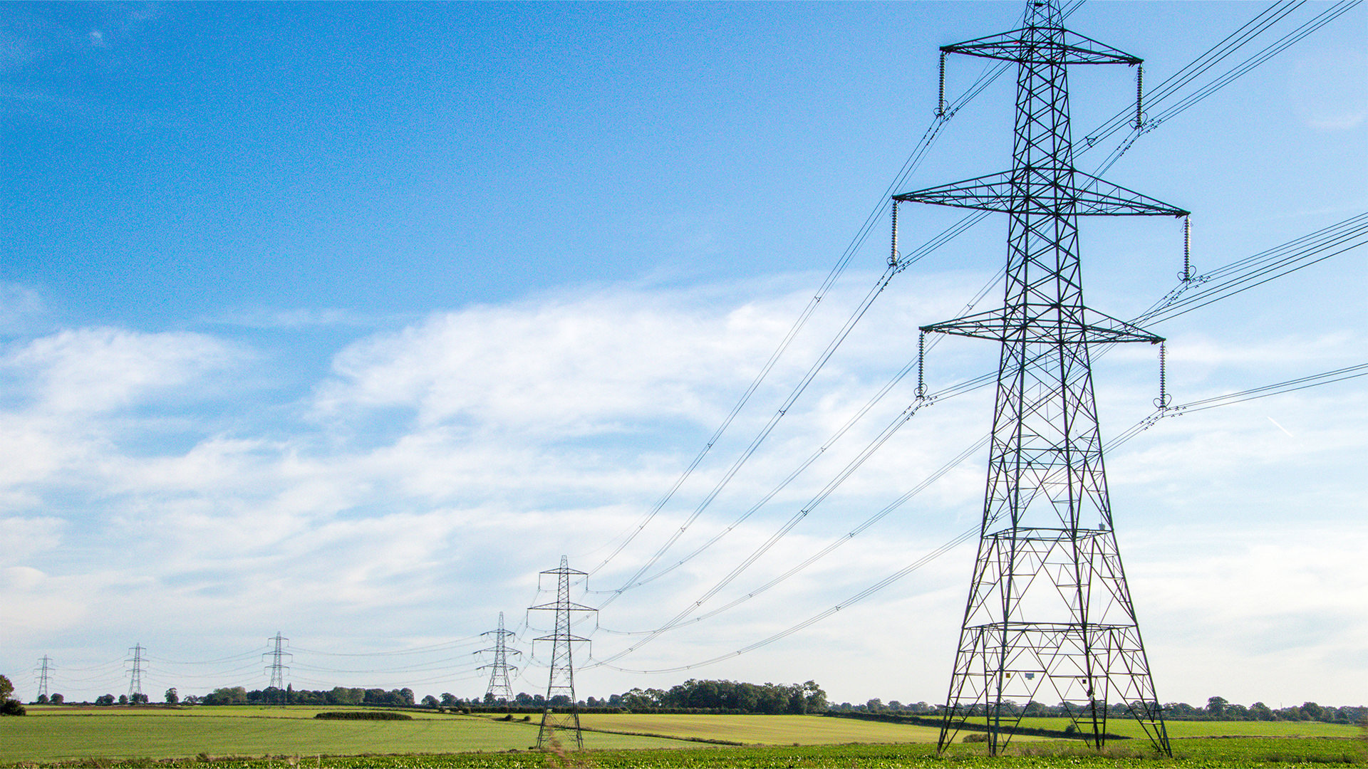 High-voltage power lines stretch across a rural landscape under a clear blue sky, with fields and trees in the background