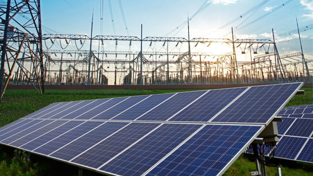 Solar panels in the foreground with a large electrical substation and transmission lines in the background, set against a bright sky with the sun partially visible, symbolizing the intersection of renewable energy and grid infrastructure