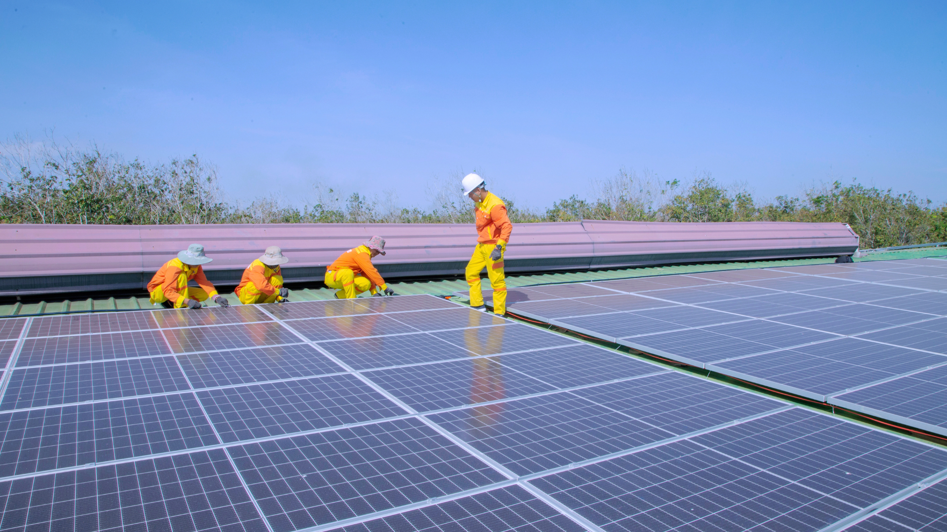 Four workers in orange uniforms are installing solar panels on a roof, carefully positioning and securing them under a clear blue sky, illustrating the process of solar panel installation on a commercial or industrial scale.