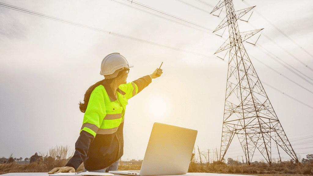 A utility worker in a high-visibility jacket and hard hat, holding a two-way radio and pointing towards a tall electrical transmission tower.