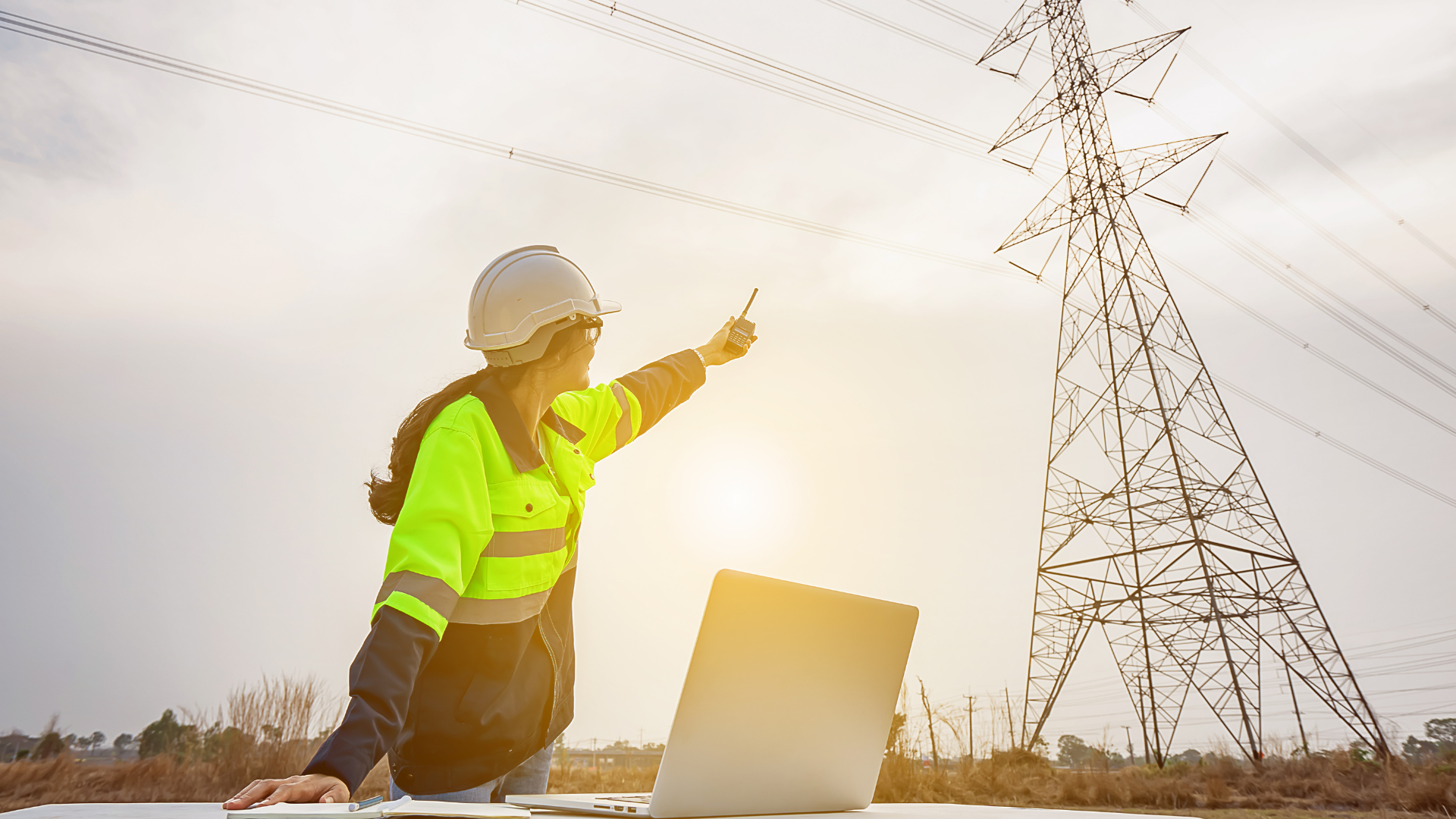 A utility worker in a high-visibility jacket and hard hat, holding a two-way radio and pointing towards a tall electrical transmission tower.