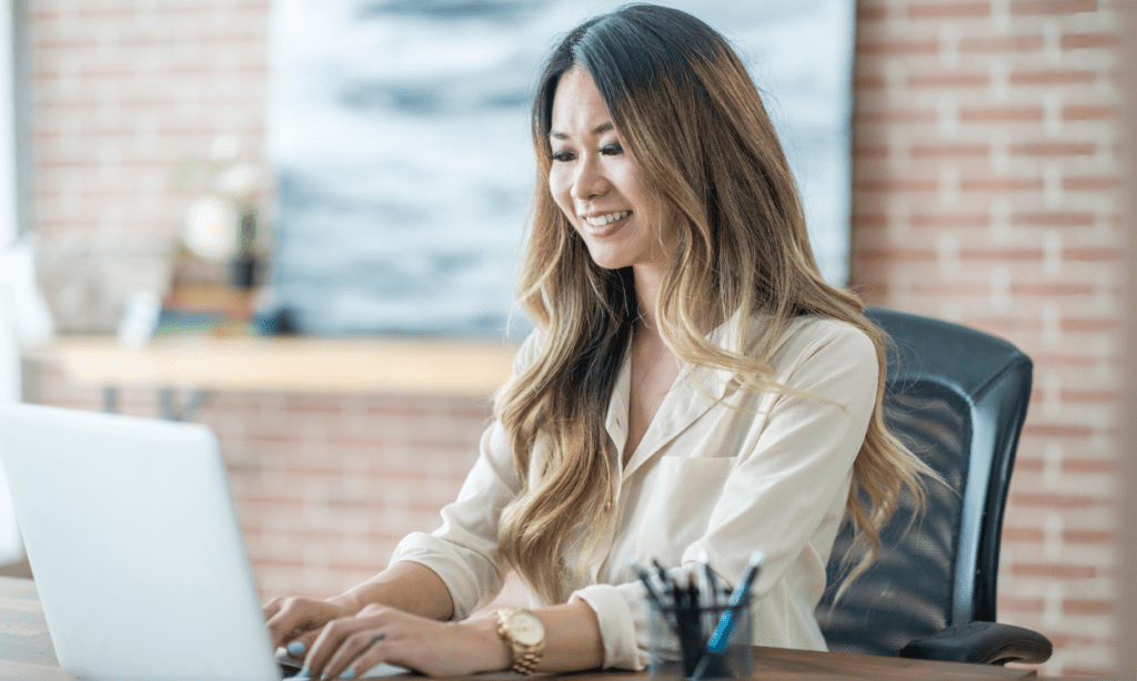 Smiling woman sitting at a desk, working on a laptop in a modern office setting.