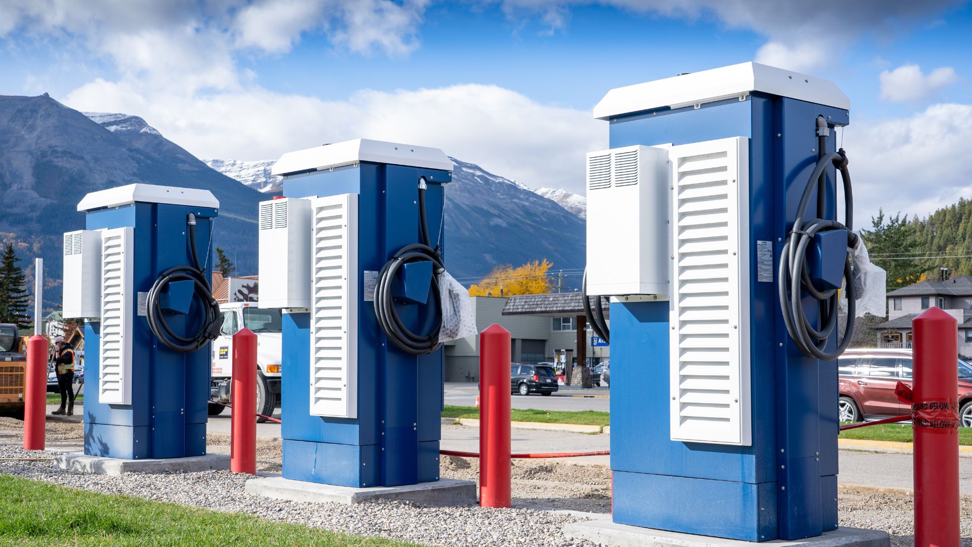 Three blue EV charging stations with attached cables in a scenic outdoor setting with mountains and a cloudy sky in the background