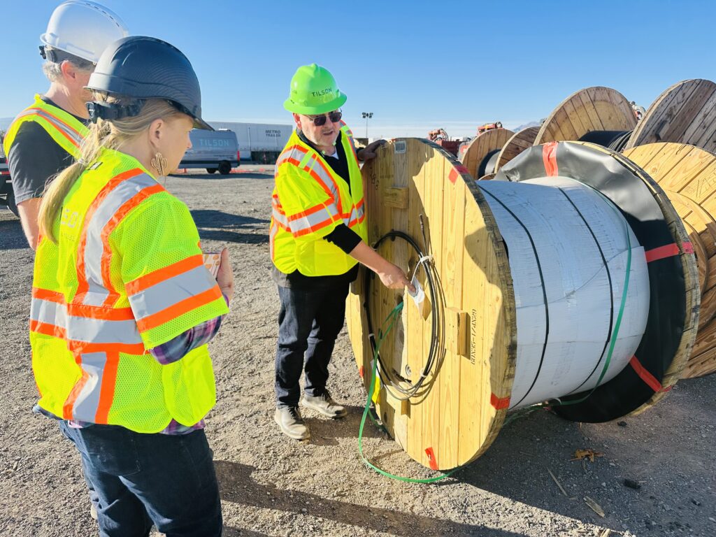 Three Tilson workers in safety gear inspecting a large wooden spool of fiber optic cable at a construction site under clear blue skies