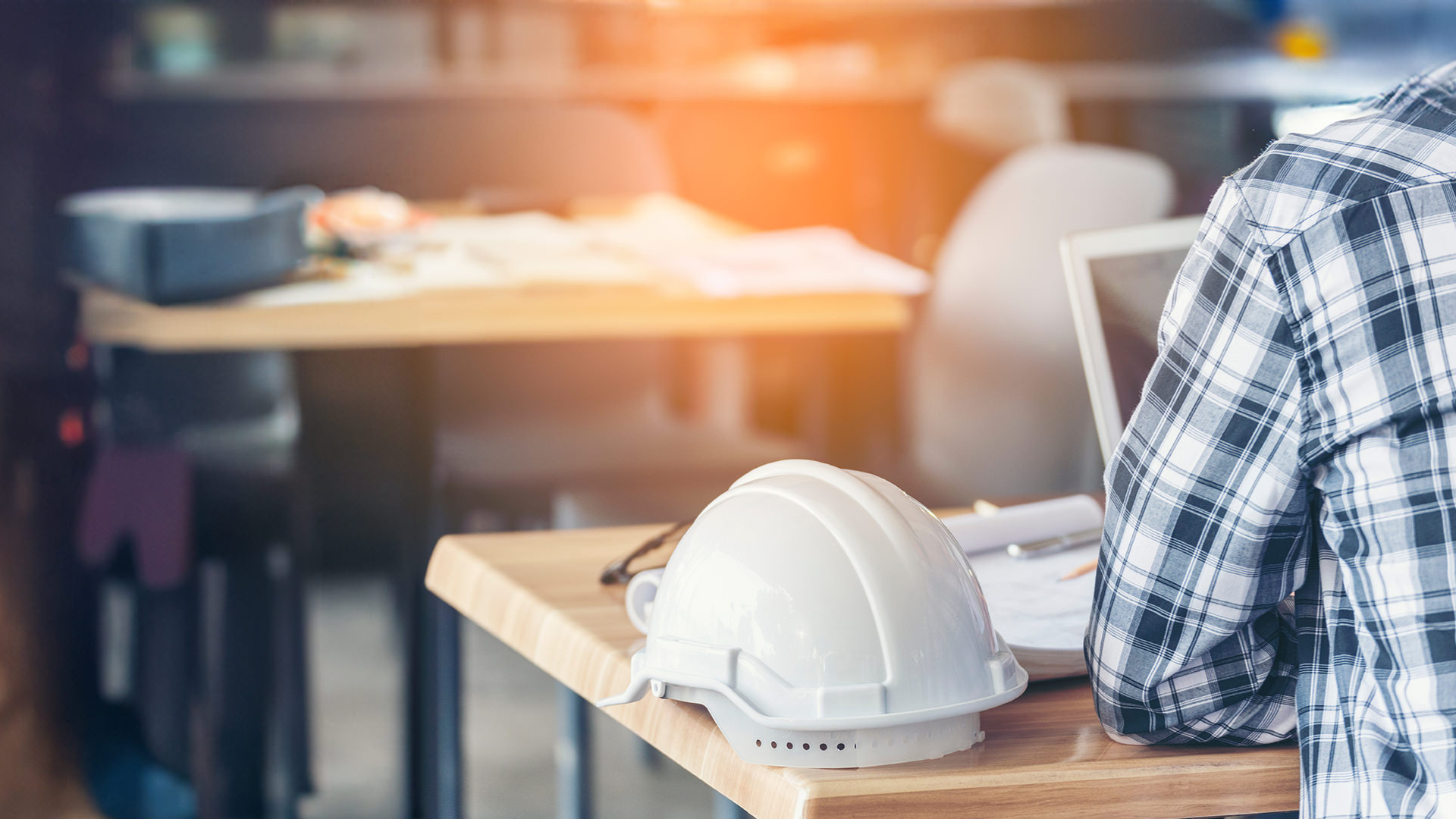 Close-up of a white hard hat on a wooden desk in a construction office, with a person in a plaid shirt working on a laptop in the background.