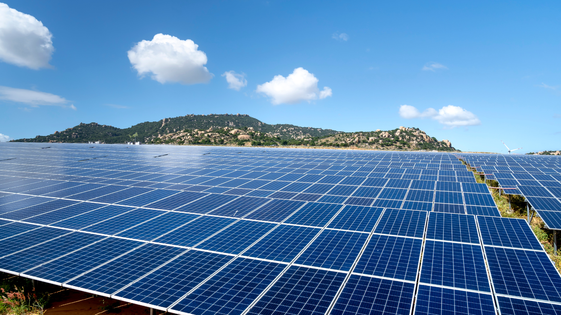 A vast solar power farm with rows of photovoltaic panels under a bright blue sky with fluffy white clouds. A green, hilly landscape is visible in the background.