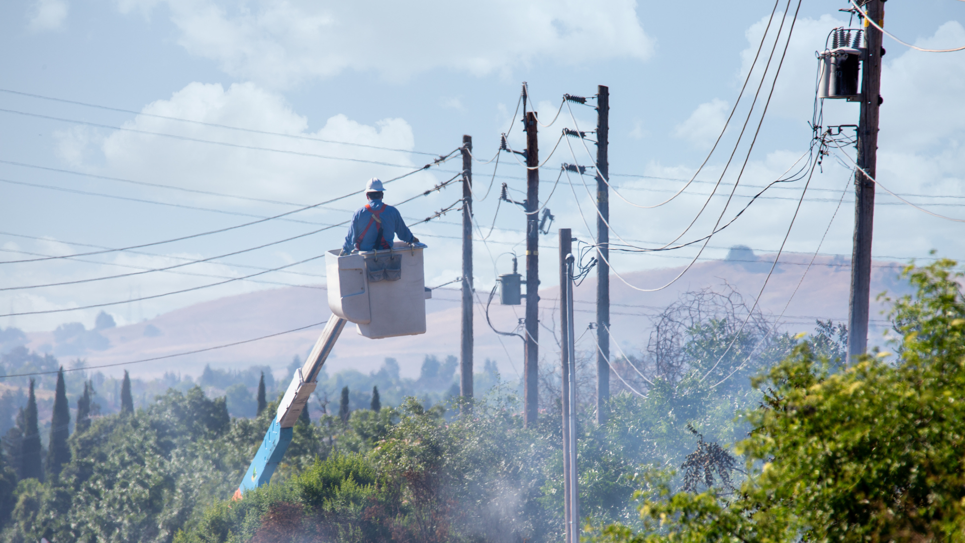 Utility worker in a bucket lift maintaining power lines, surrounded by trees and distant hills under a partly cloudy sky.