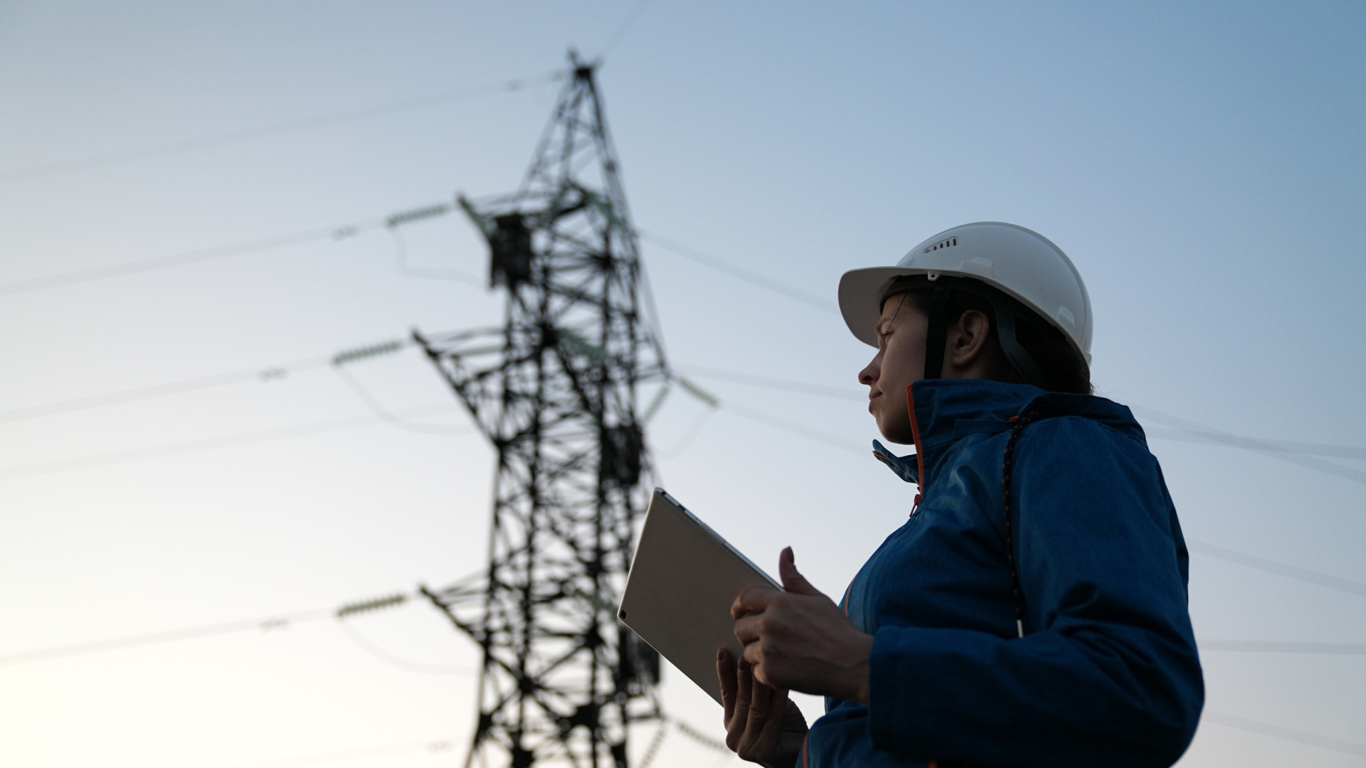 Female utility worker wearing a white hard hat and blue jacket, holding a tablet while inspecting a high-voltage power transmission tower against a clear sky at dusk